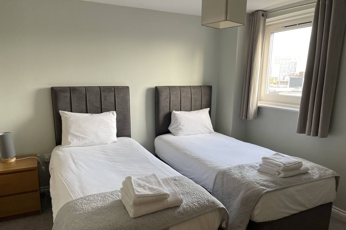 Une chambre d’hôtel avec deux lits, chacun doté d’une tête de lit grise, de draps blancs et d’une fenêtre avec des rideaux.