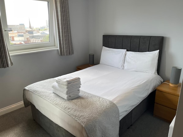 A neatly made bed with a grey headboard, white pillows, and a folded towel on the footboard. There is a bedside lamp on a wooden nightstand beside the bed.