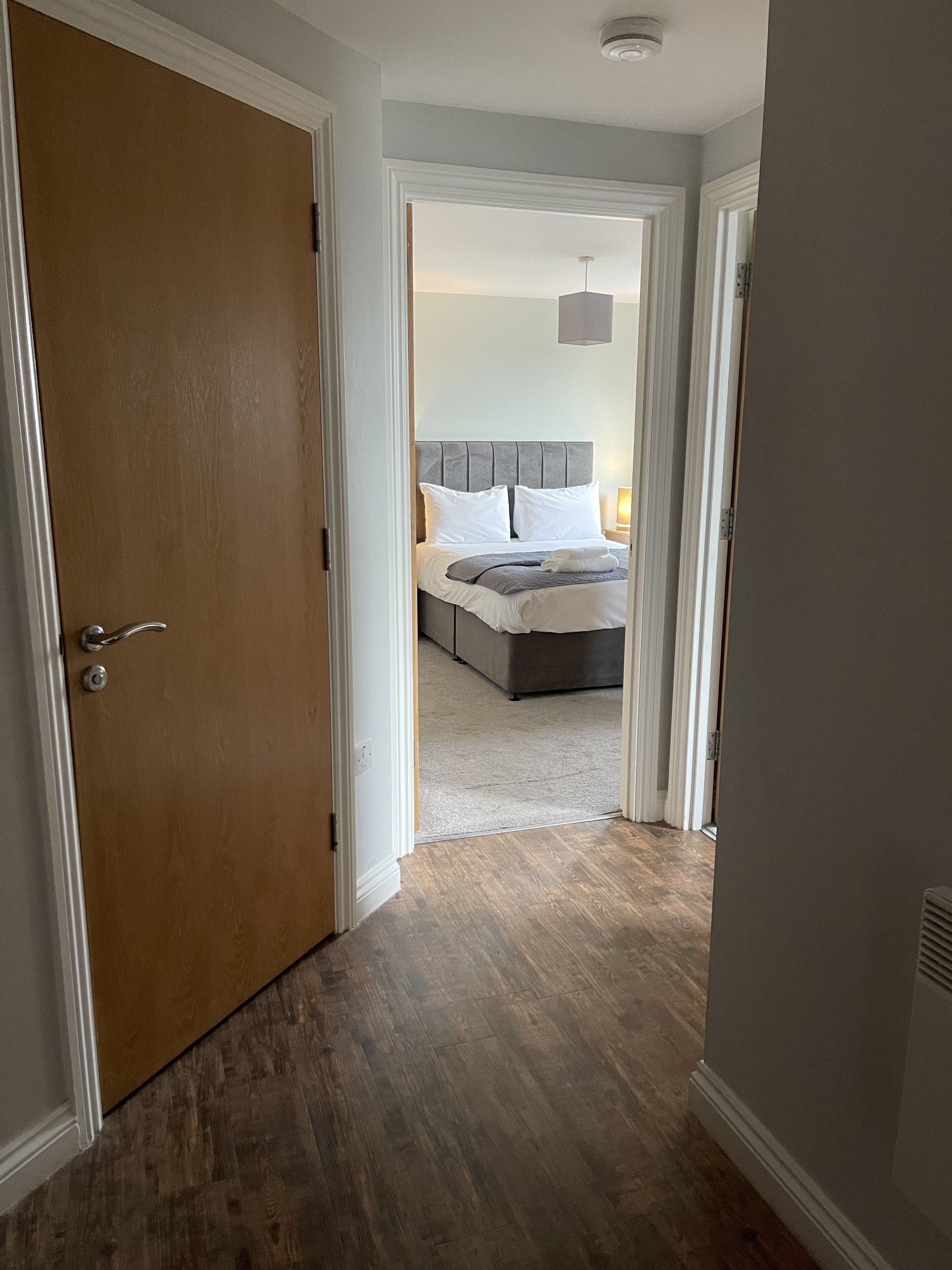 Un couloir menant à une chambre à coucher équipée d’un lit, d’une table de chevet et d’un luminaire au plafond.
