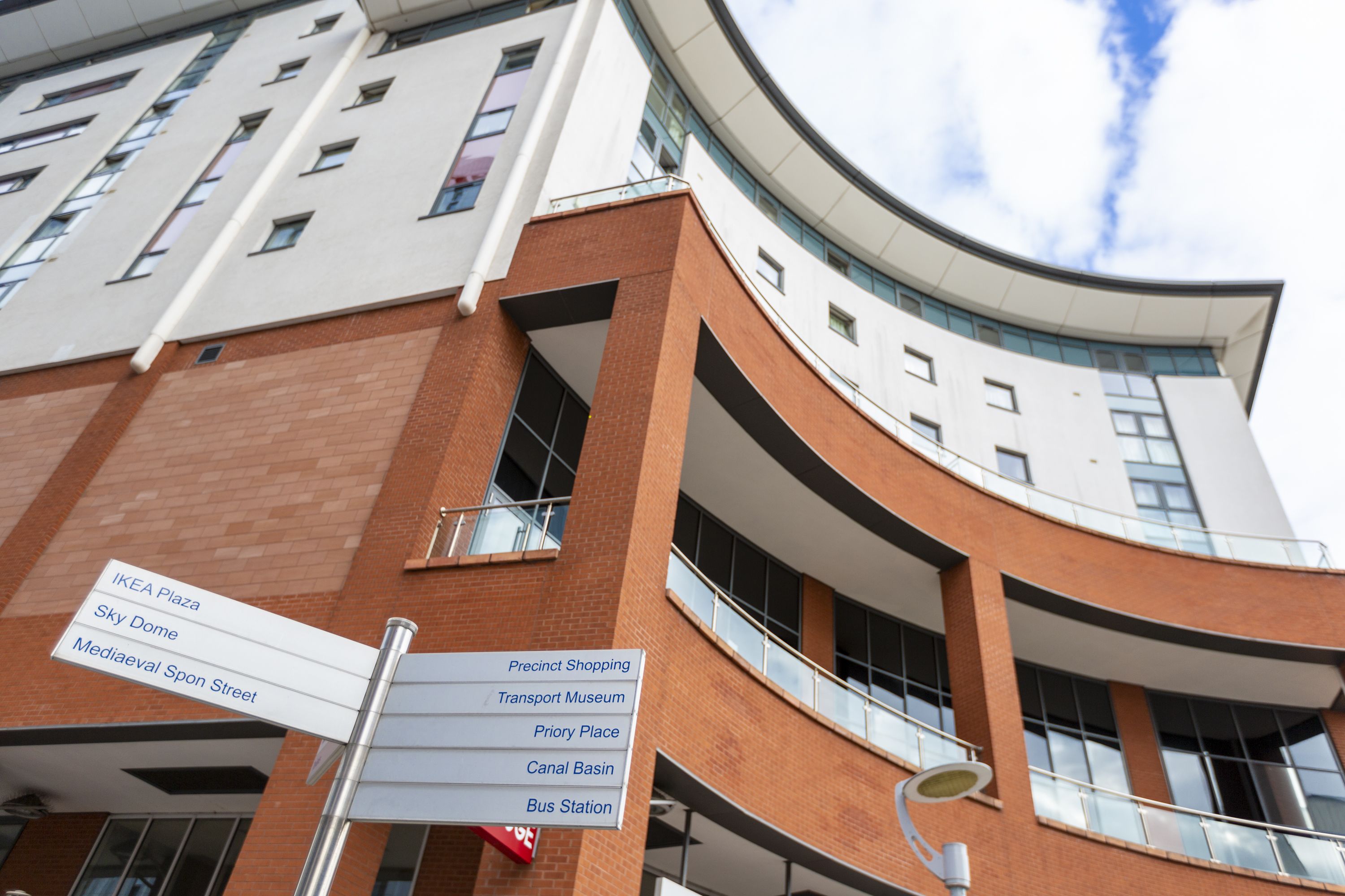 Egy modern épület, ívelt homlokzattal, melyen táblák jelzik az irányt a különböző helyszínek felé, többek között az IKEA Plaza, a Sky Dome és a Mediaeval Spon Street felé.