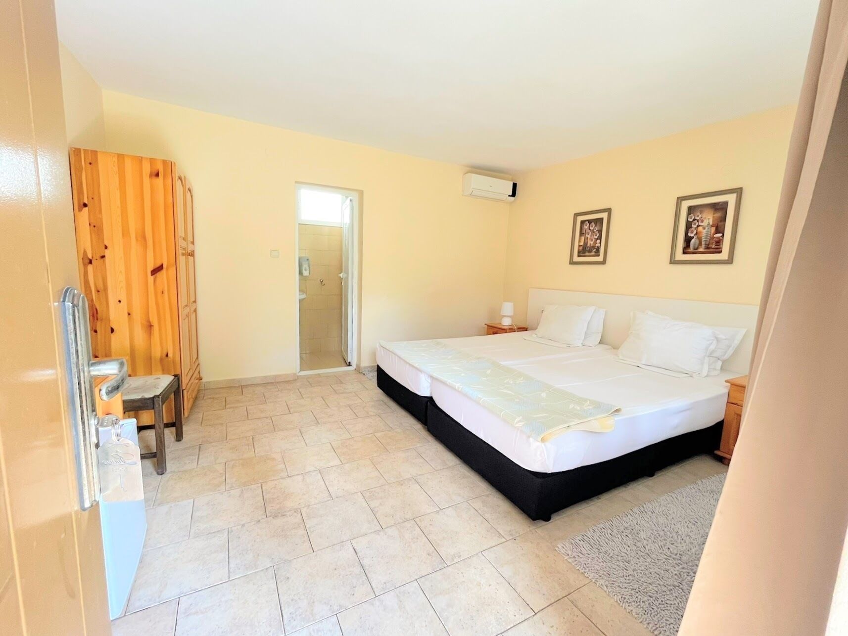 A bedroom with a bed, a wooden wardrobe, a small table, a nightstand, and a door leading to a bathroom.