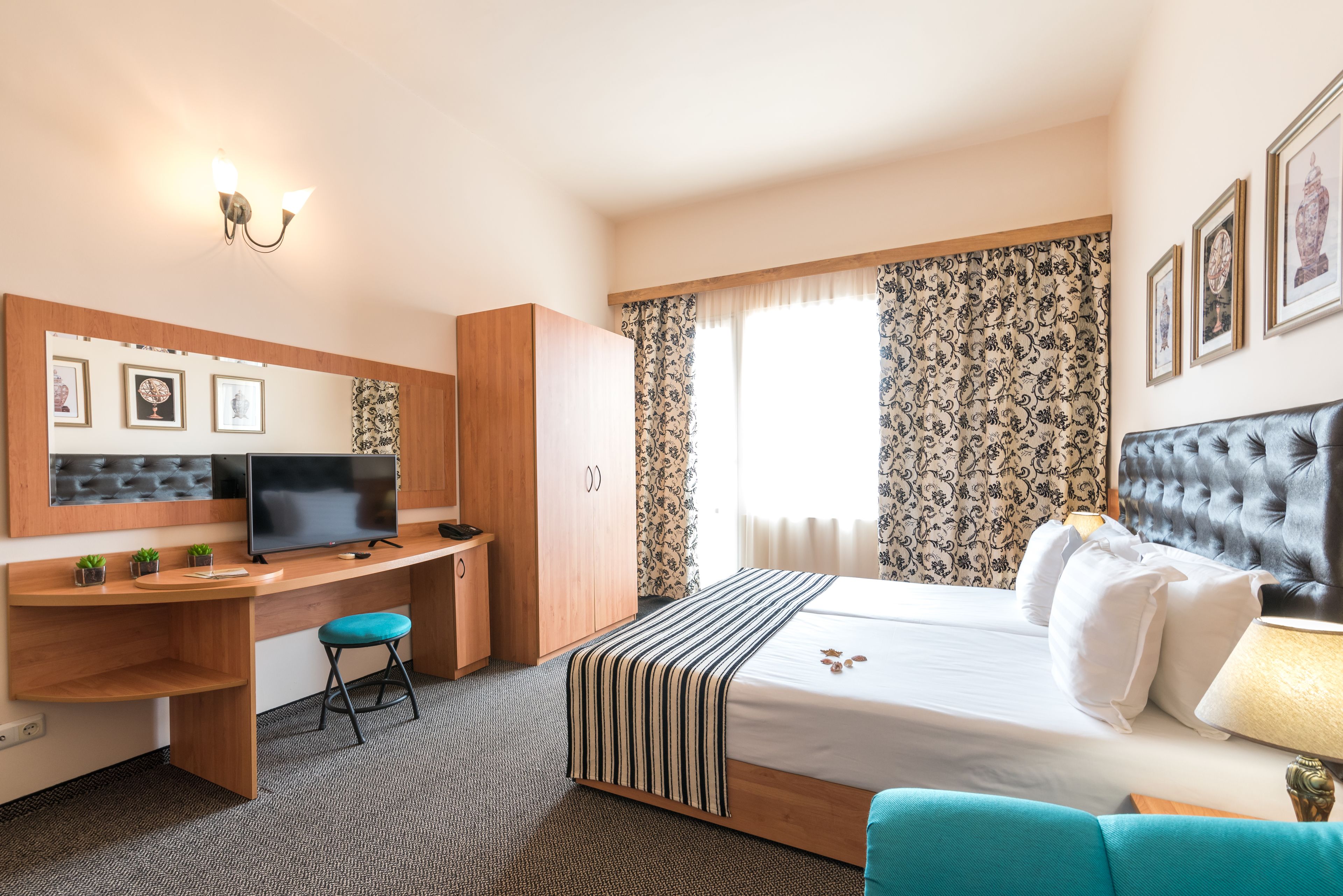 A hotel room with a bed, desk, and chair, a television, and a window with patterned curtains.