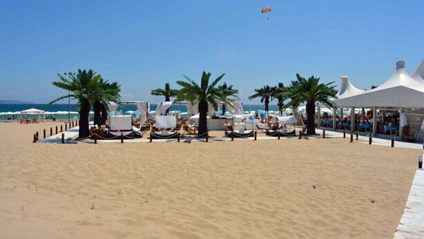 Un espace aménagé au bord de la plage, avec des chaises longues, des palmiers et une structure blanche tendue en forme de tente.