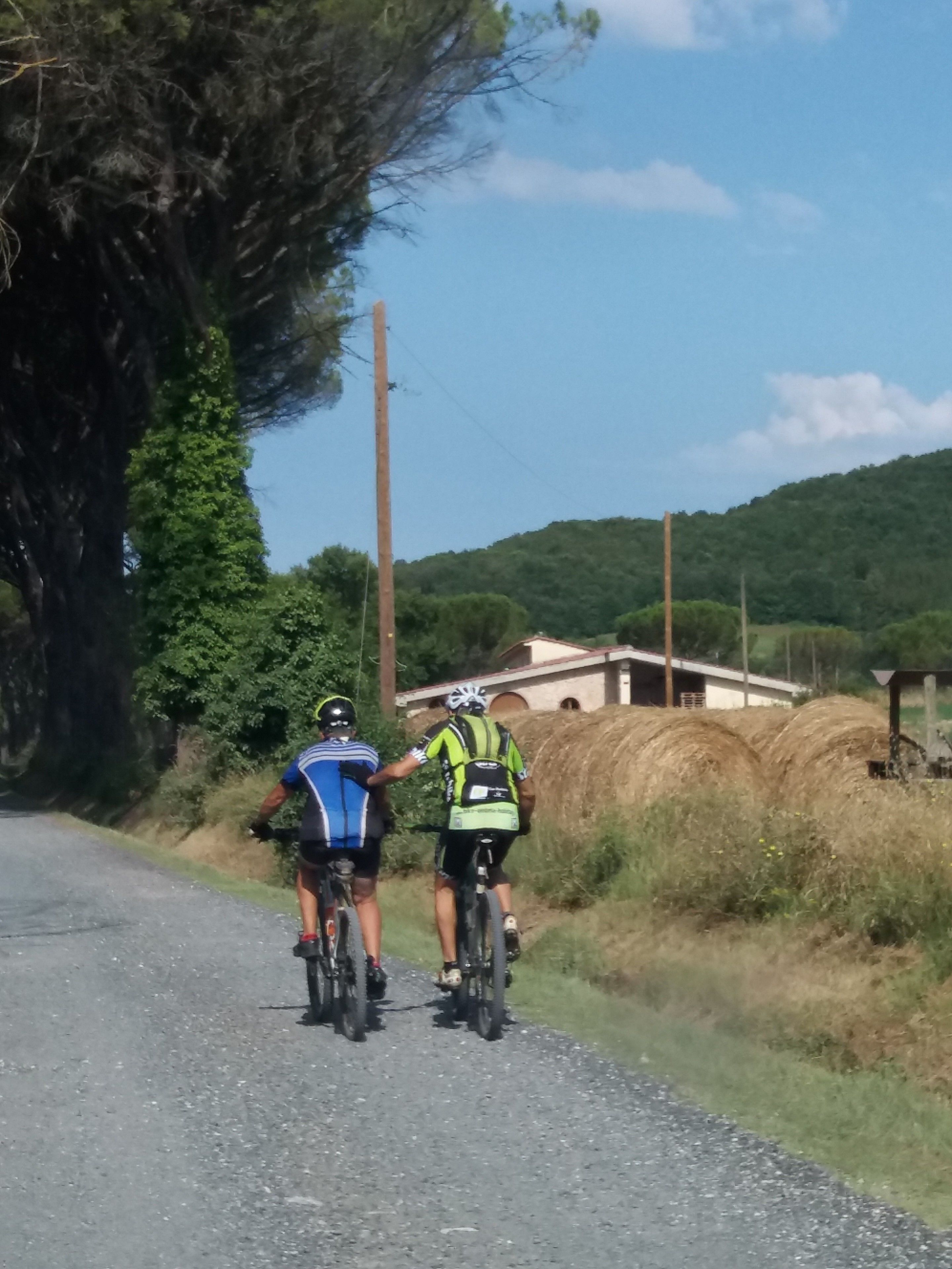 Deux cyclistes sur un chemin de terre, avec des balles de foin et un bâtiment en arrière-plan.