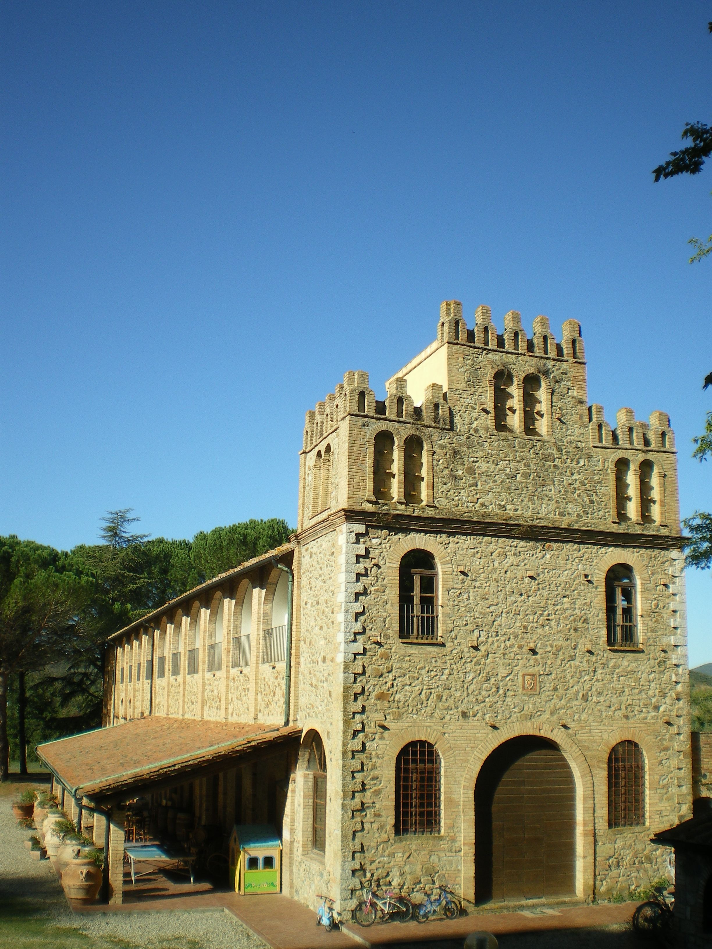 Un edificio storico in pietra con una torre, finestre ad arco e un portico coperto con piante in vaso.