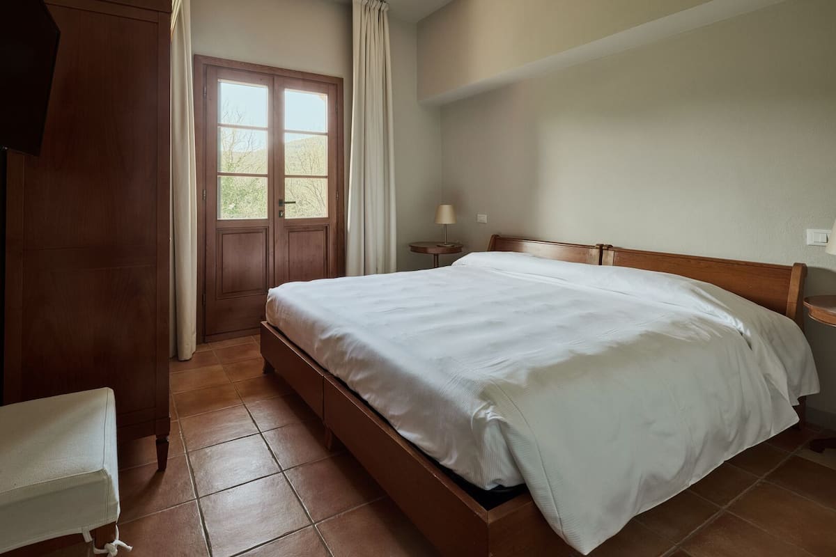 Un dormitorio con una cama de madera, una mesita de noche, una lámpara y vistas al exterior a través de una puerta.