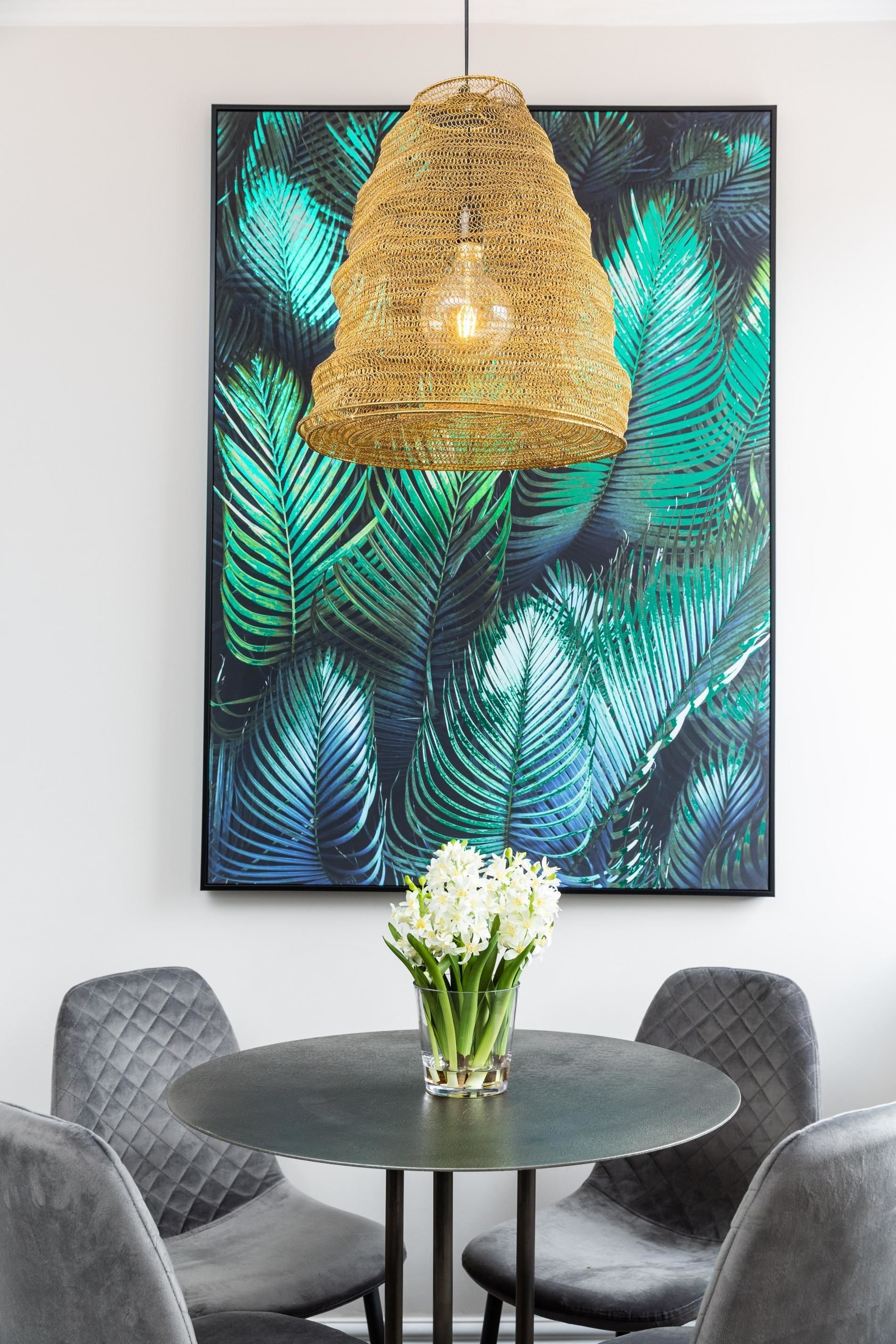 A round table with a vase of white flowers, surrounded by grey upholstered chairs, under a woven pendant light, with a tropical leaf pattern artwork on the wall behind.