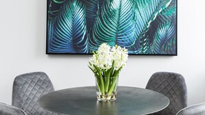 Una mesa redonda con un florero de flores blancas, rodeada de sillas tapizadas en gris, bajo una lámpara colgante de fibra, con un cuadro en la pared detrás que presenta un motivo de hojas tropicales.