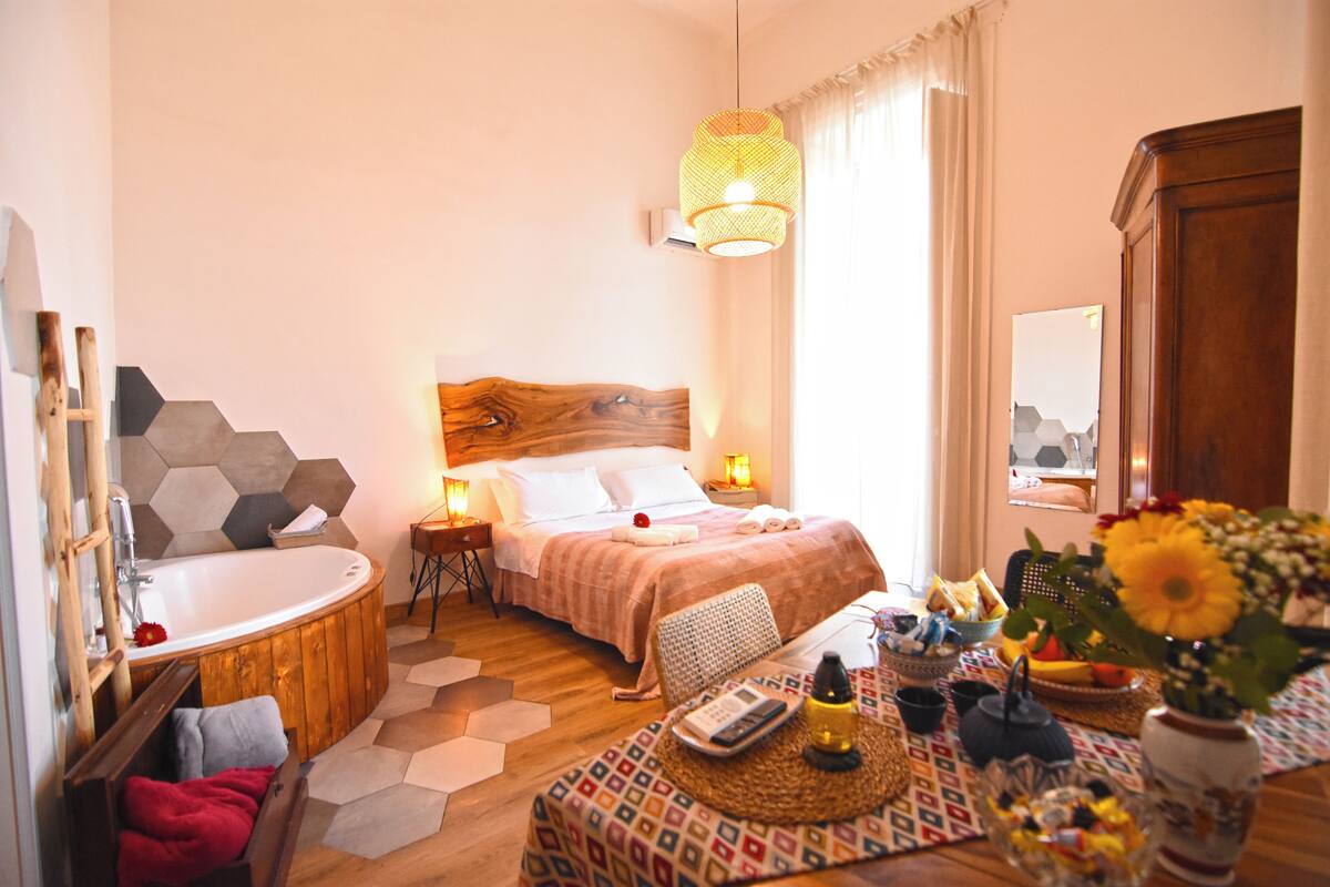 A modern bedroom with a wooden headboard, a large bed, a wooden bathtub, a bedside table with a lamp, and a small table with a vase of flowers.