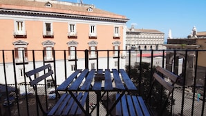 A balcony with a wooden table and chairs, overlooking a street with buildings and a statue. - Sunrise Luxury (Naples)