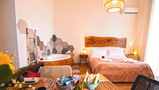 A hotel room with a wooden headboard, a wooden side table, a round wooden table with fruits and towels, a wicker chair, and a wall-mounted air conditioner. - Sunrise Luxury (Naples)