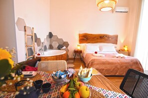 A hotel room with a wooden headboard, a wooden side table, a round wooden table with fruits and towels, a wicker chair, and a wall-mounted air conditioner. - Sunrise Luxury (Naples)