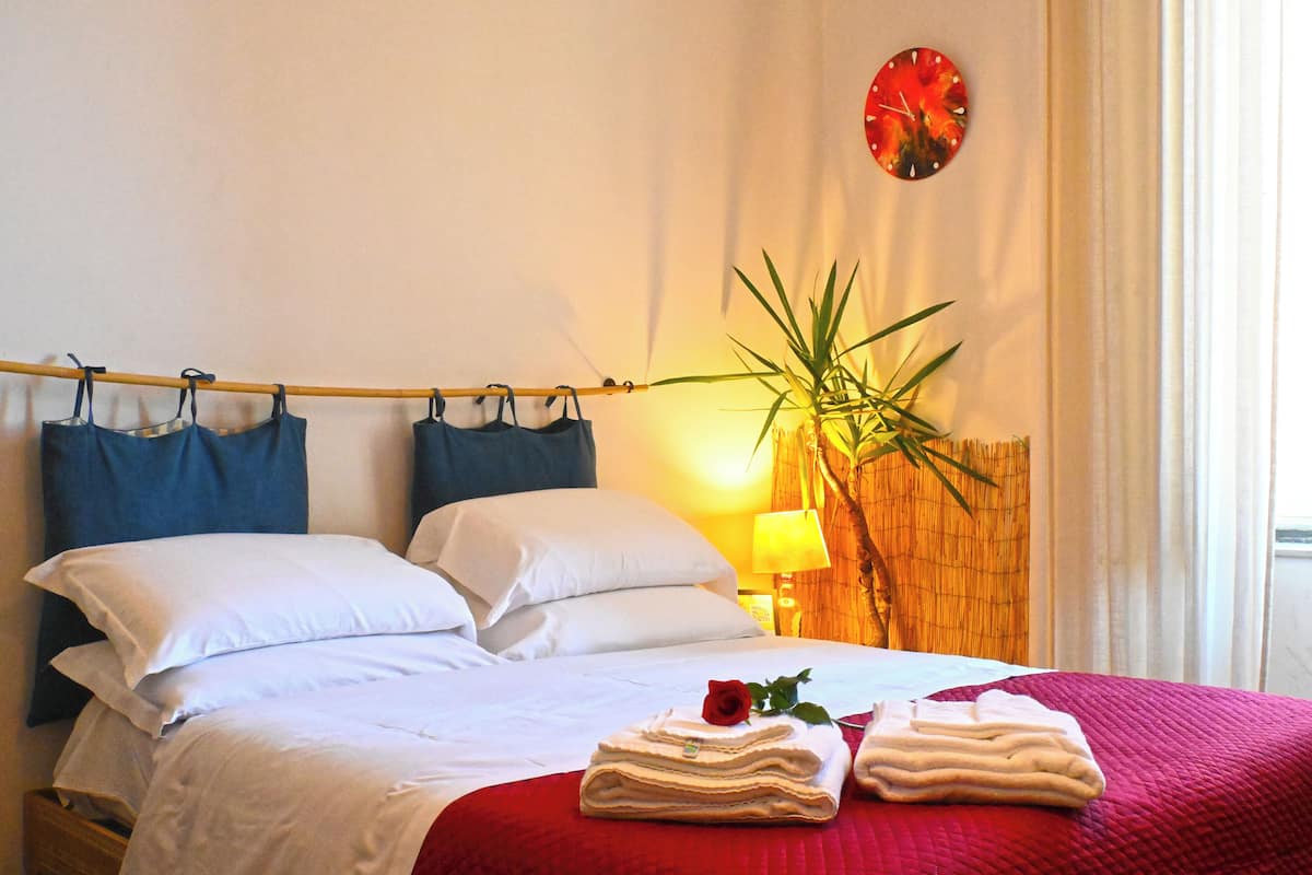 A neatly made bed with white linens, a red blanket, and a rose on the pillows.
