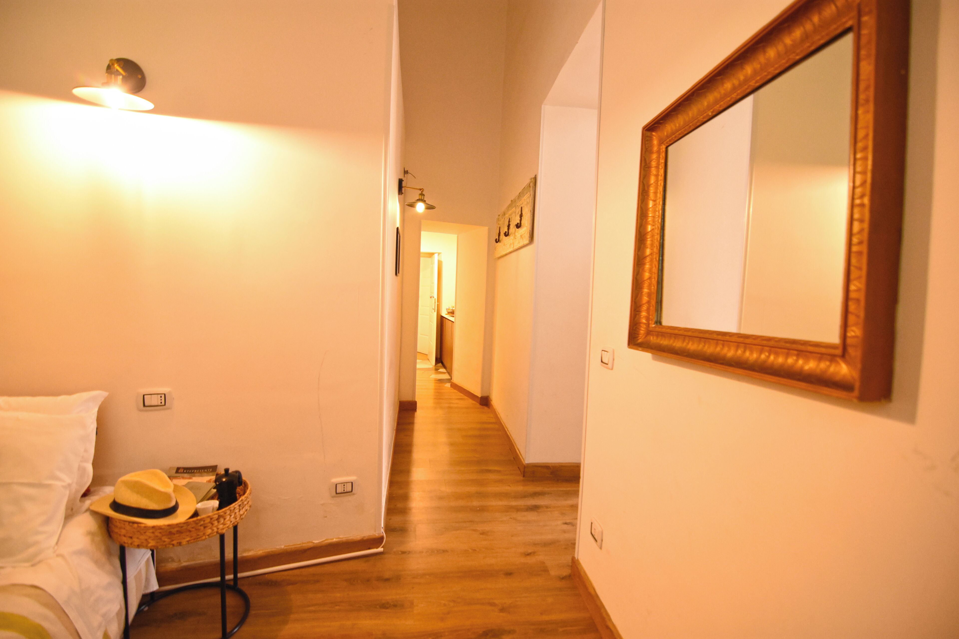 Une chambre d’hôtel avec un lit, une table de chevet sur laquelle se trouvent un chapeau et un appareil photo, un miroir, et un couloir menant à une autre pièce.