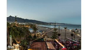 Ein Küstenort mit Strand, einem Berg im Hintergrund und einer Bar mit Menschen.