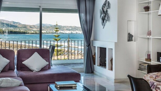 Ein modernes Wohnzimmer mit großem Fenster, das Blick auf Strand und Berge bietet, ein violettes Sofa und ein Kamin.