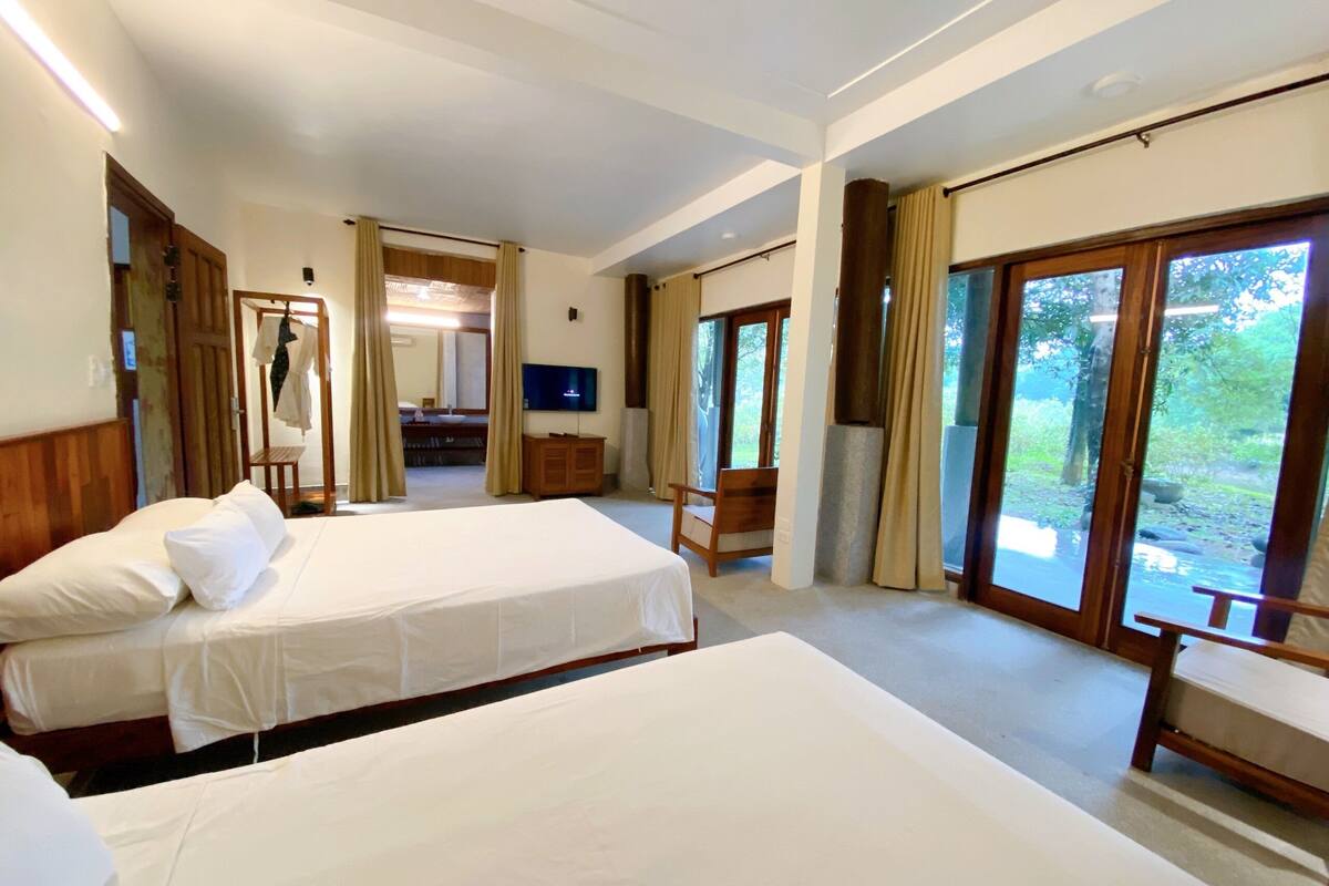 A hotel room with two beds, a TV, a desk, and large windows.