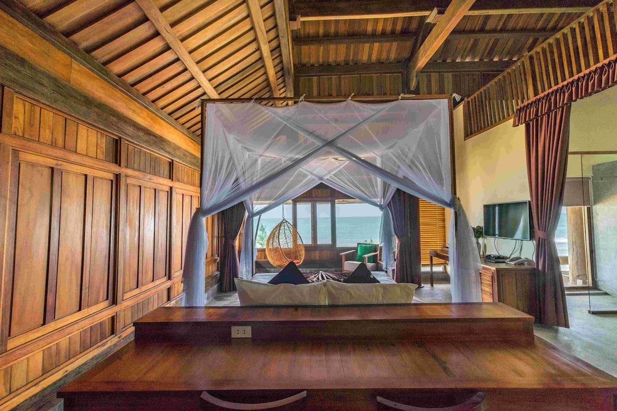 A room with a bed under a mosquito net, wooden walls, and a view of the sea.