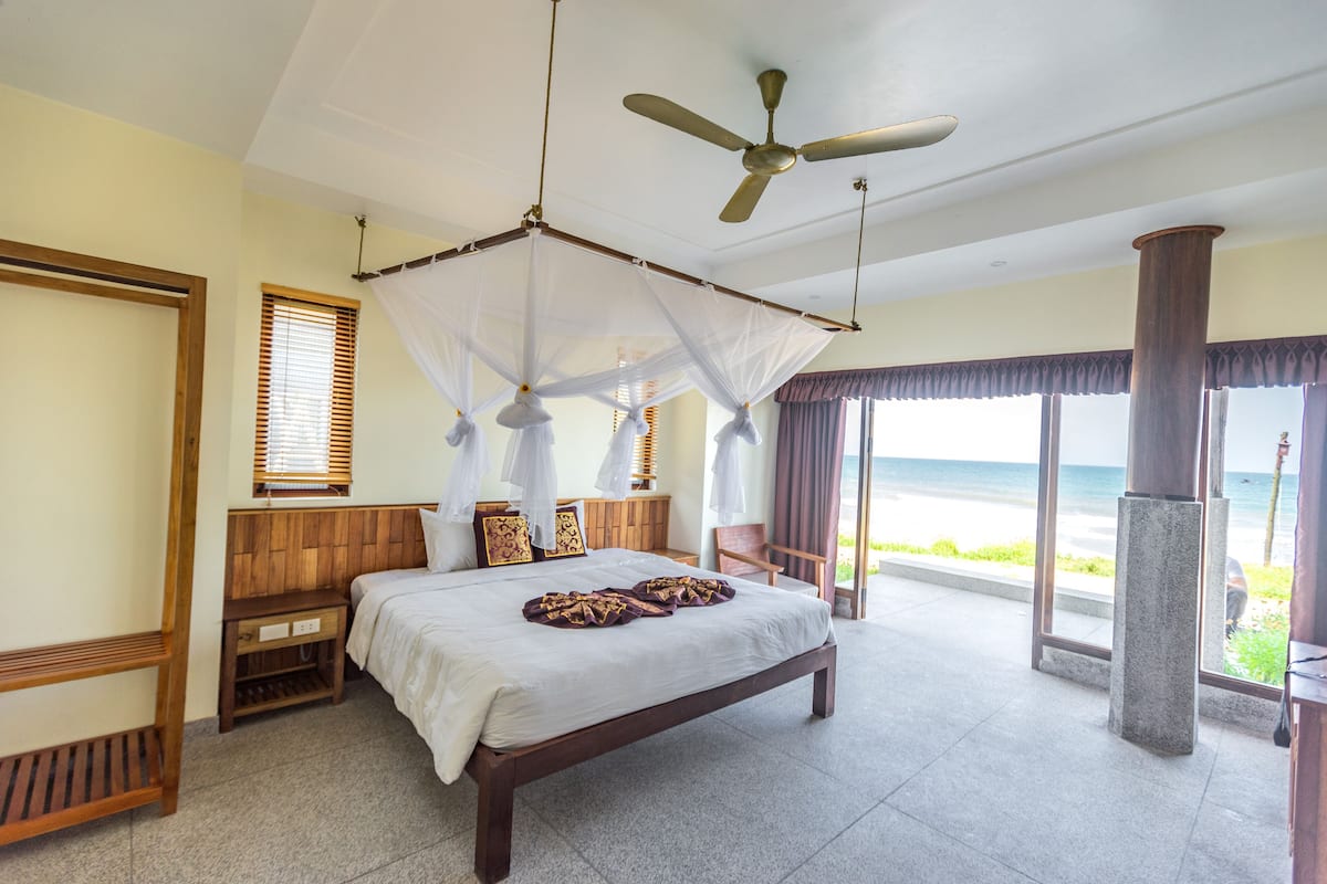 A bedroom with a bed, wooden headboard, bedside tables, a ceiling fan, and a view of the ocean.