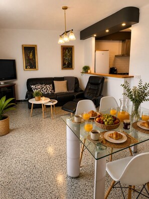 A modern living room with a glass dining table, a sofa, a TV, and a kitchen area. - Ap. NURIMAR 2 (In the Center of the Village) (La Escala)
