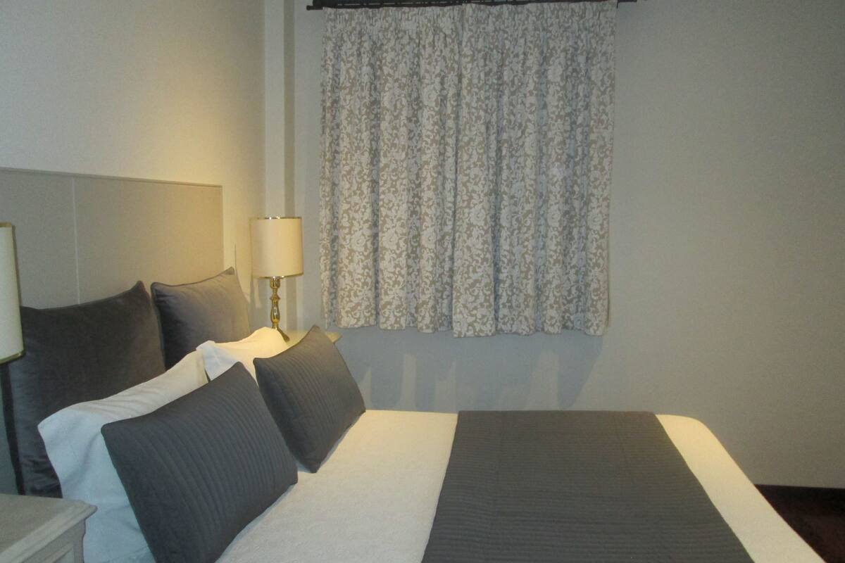 Een net opgemaakt bed met een deken in donker- en lichtstrepen, twee kussens, een bedlampje en een gordijn met bloemenprint.