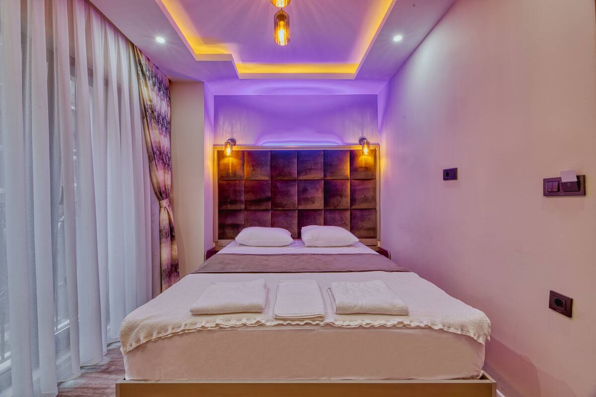 A modern bedroom with a large bed, a headboard with a textured panel, and ambient lighting.