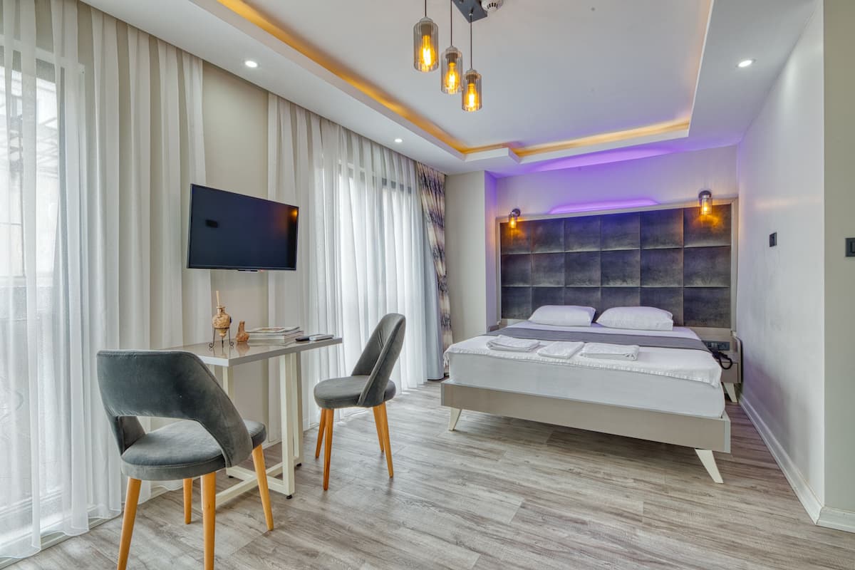 A modern hotel room with a large bed, a desk with a flat-screen TV, two chairs, and a large window with sheer curtains.