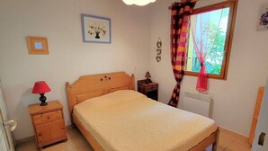 Un dormitorio con cama de madera, mesita de noche con lámpara, ventana con cortinas y radiador.