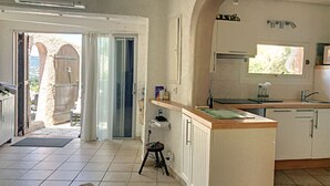 Een keuken met een centraal eiland, een eethoek met een tafel en stoelen, en uitzicht door een open deur op een buitenruimte.