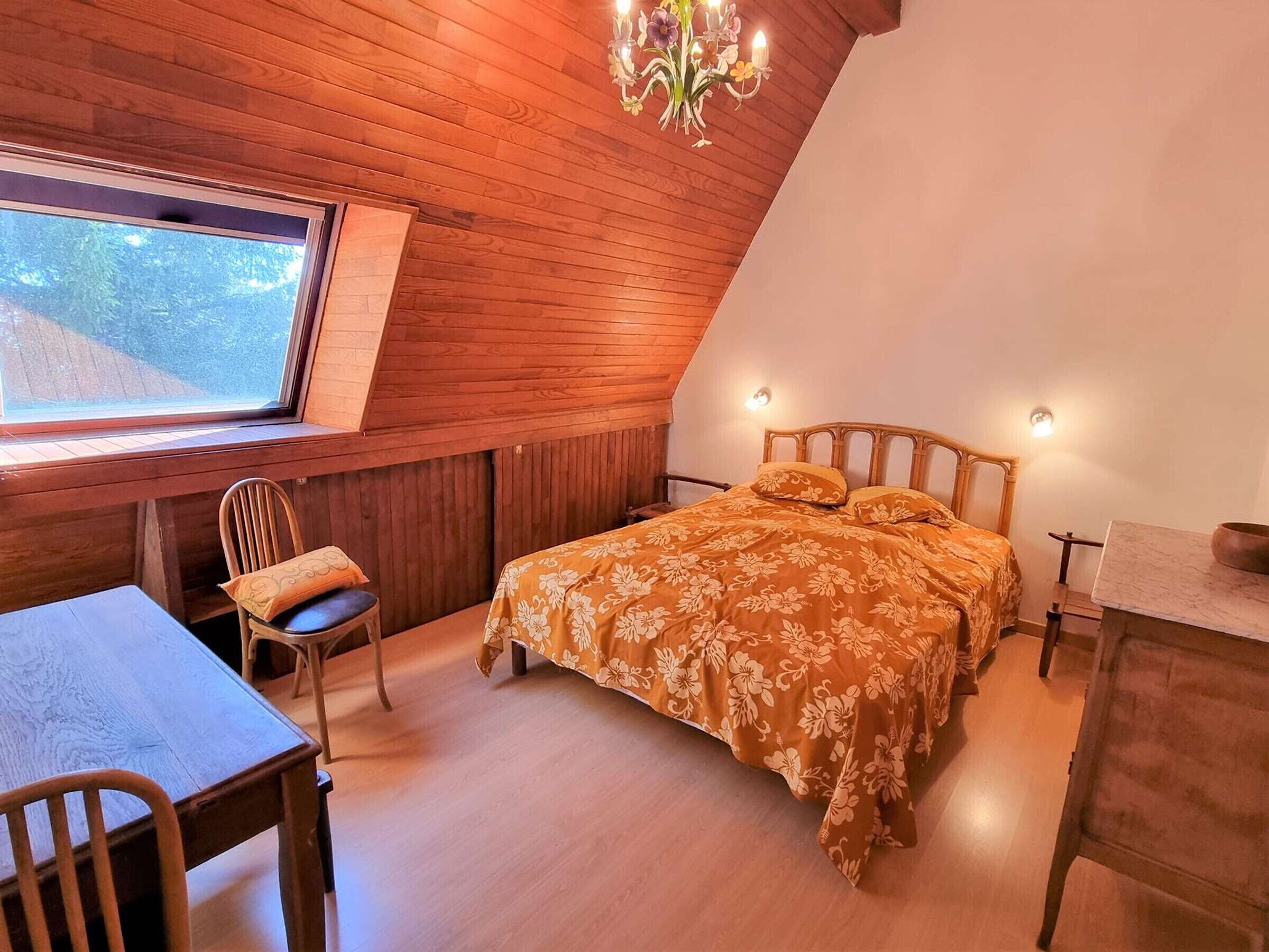 Un dormitorio acogedor con techo de madera, una cama con ropa de cama floral, un escritorio y una ventana con vistas a la vegetación.