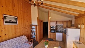 L’intérieur chaleureux d’un chalet avec un canapé en bois, une table à manger avec des chaises, un coin cuisine équipé d’appareils électroménagers et une bibliothèque.