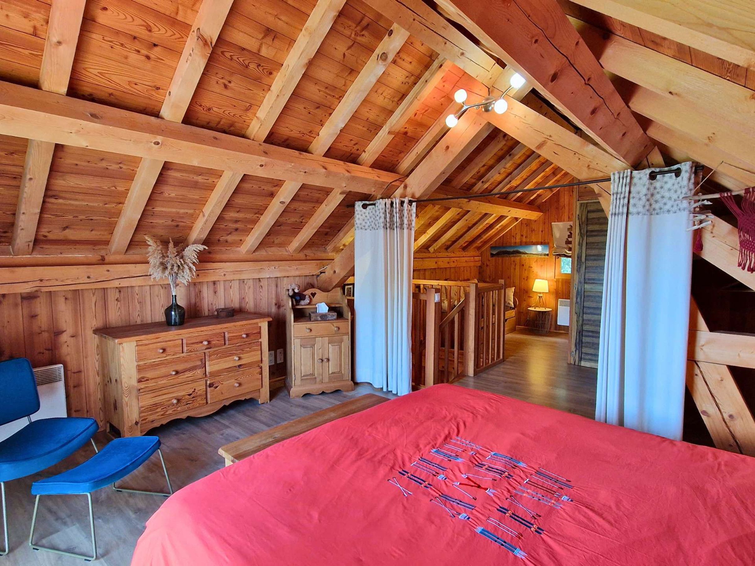 Un dormitorio acogedor en el ático con vigas de madera, una cama con colcha roja, un aparador y una silla.