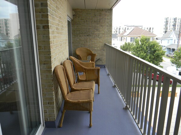 Un balcon avec des fauteuils en osier, une porte vitrée et une vue sur les immeubles voisins.