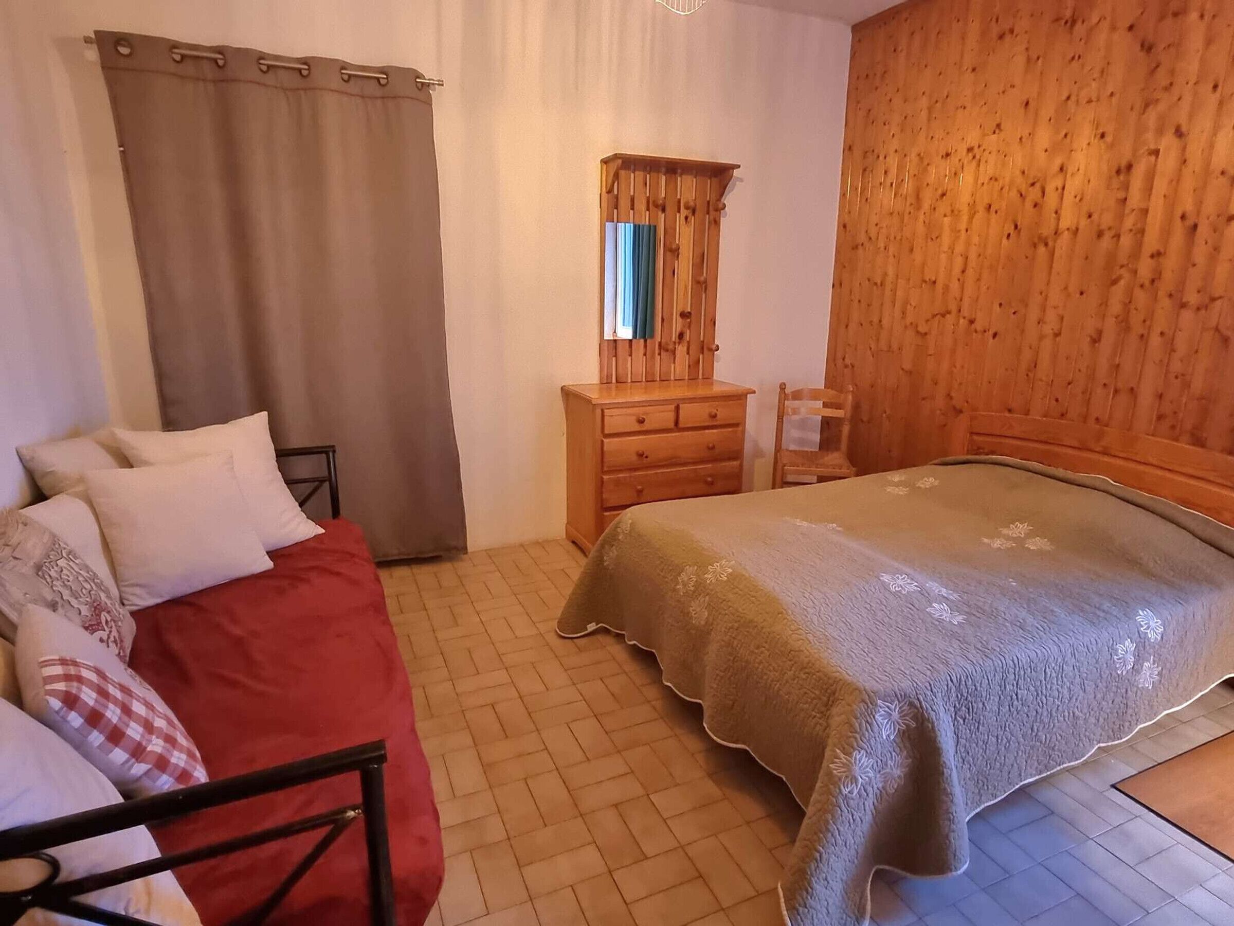 A bedroom with a bed, a wooden dresser, a mirror, and a window with curtains.