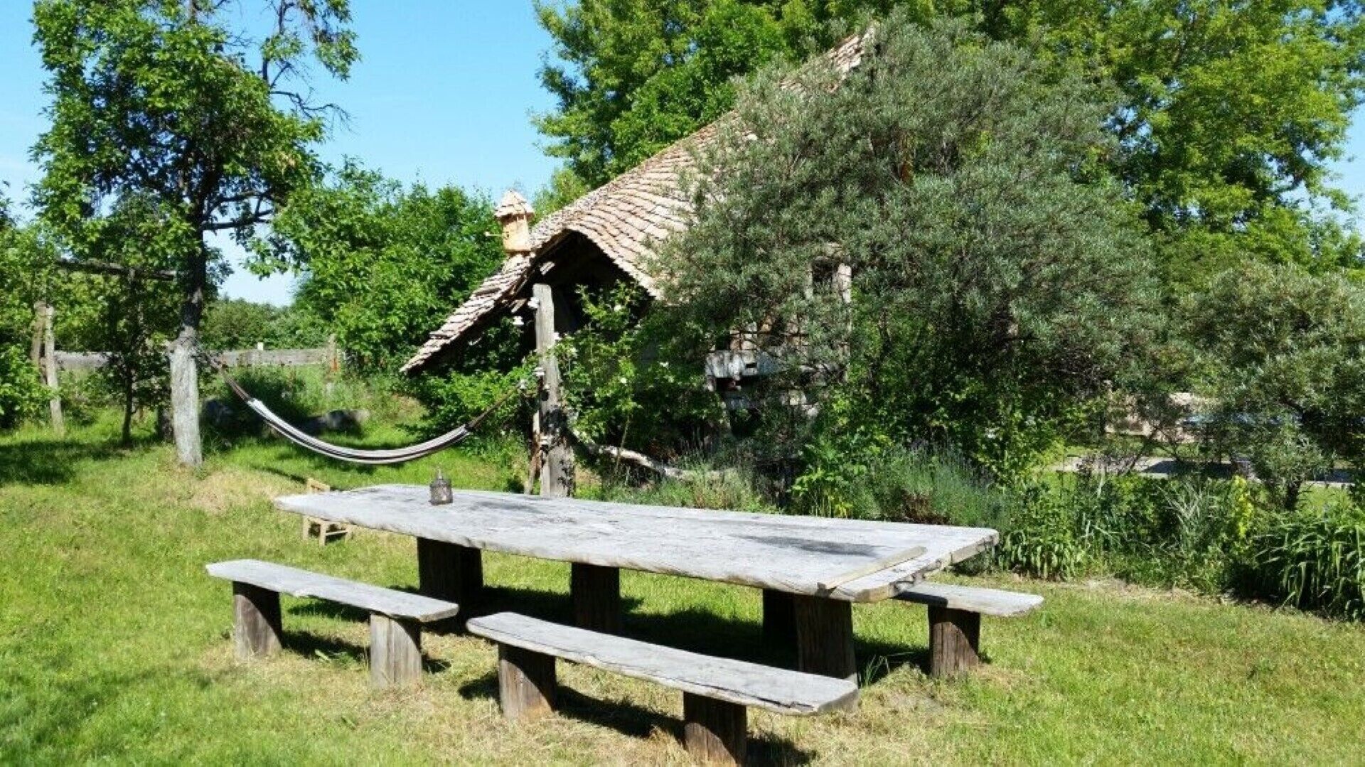 A rustic picnic table with benches, a hammock, and a small shelter with a tiled roof in a grassy area with trees.