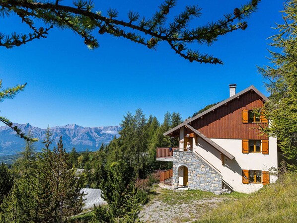 A mountain cabin with a stone staircase, wooden balcony, and a view of distant mountains.