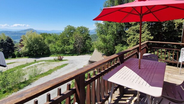 Une terrasse en bois avec une table et des chaises, un parasol rouge et une vue sur les arbres et les montagnes.