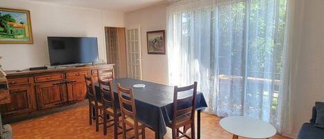 A dining area with a table and chairs, a television, and a view of greenery through large windows.