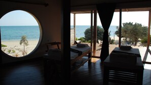 Una habitación con ventanal circular con vistas a una playa, una cama, una mesita y un banco.