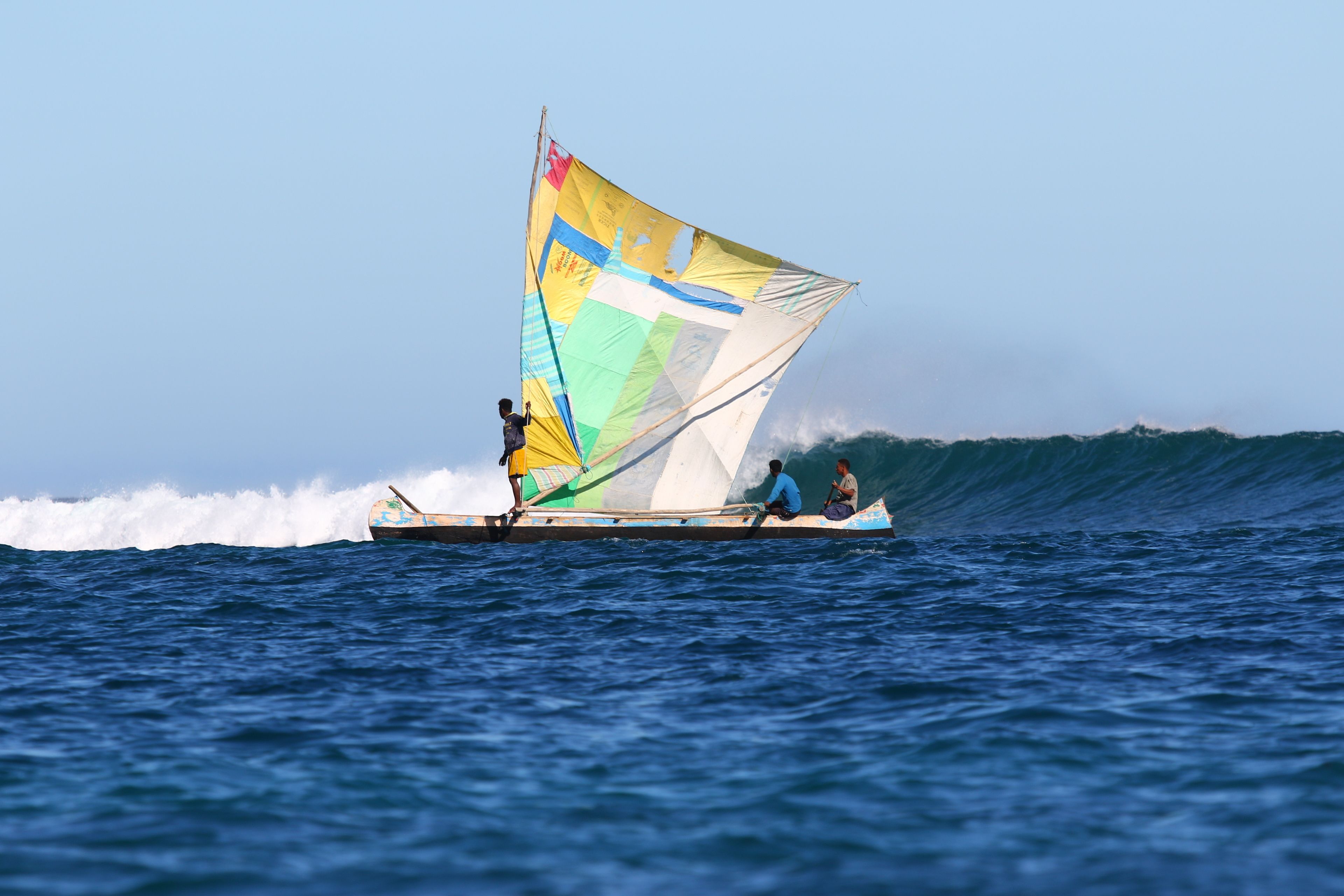 A traditional sailboat with a colorful sail, two people on board, and a person standing at the stern.