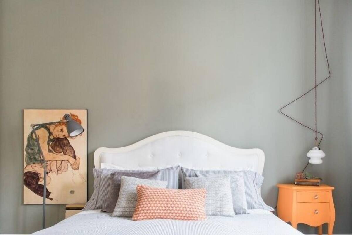 A neatly made bed with a white headboard, multiple pillows, and a bedside table with a lamp. A painting hangs on the wall beside the bed.