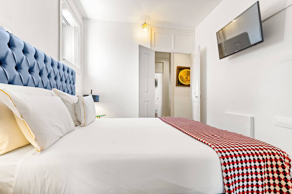 Een net opgemaakt bed met wit en crème beddengoed, een blauw gestuikt hoofdbord en een rood-zwarte geruite plaid.