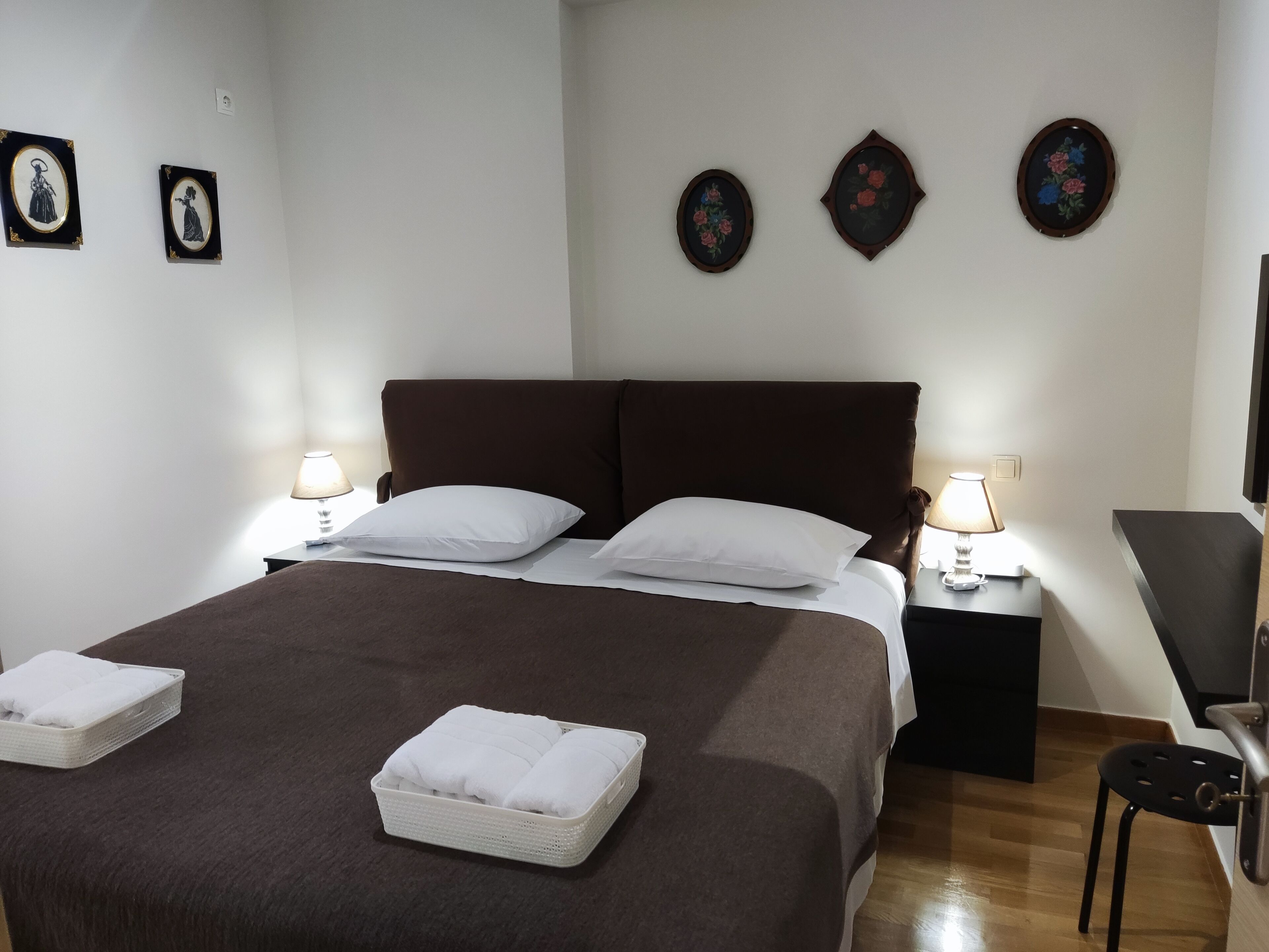 Una cama bien hecha con sábanas blancas y dos almohadas, una mesita de noche con lámpara y una pequeña silla.
