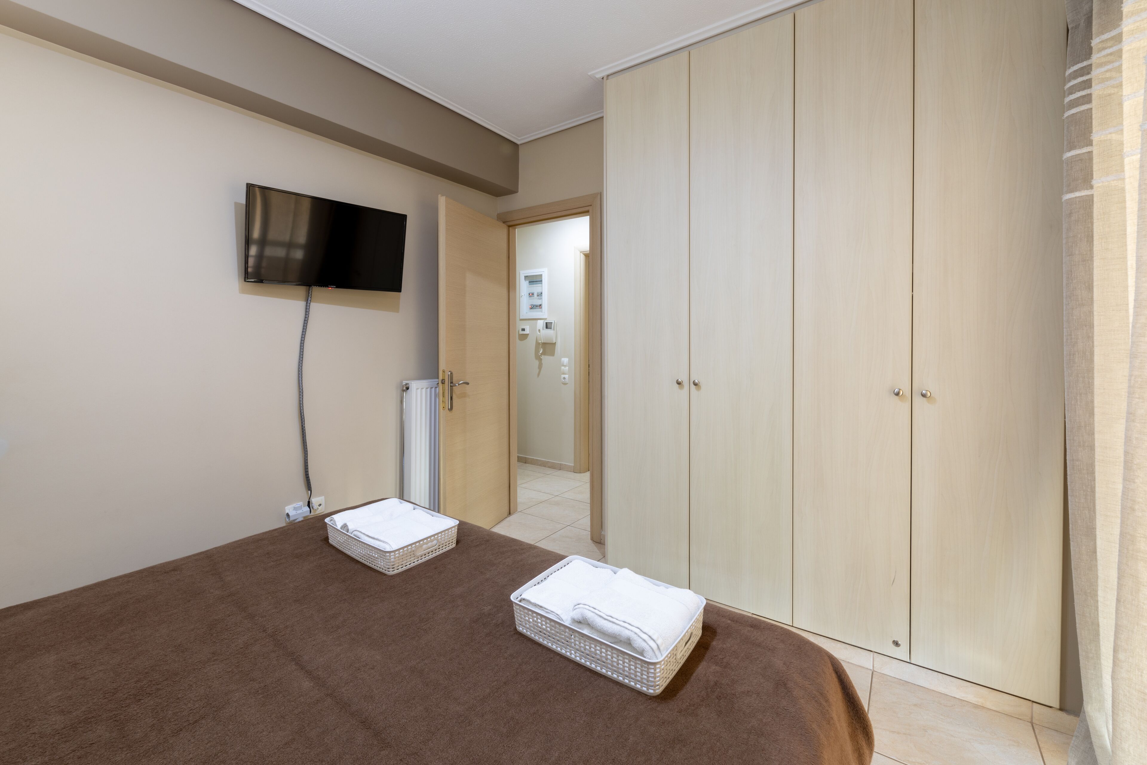 A bedroom with a large wardrobe, a flat-screen TV mounted on the wall, and two white laundry baskets on the bed.