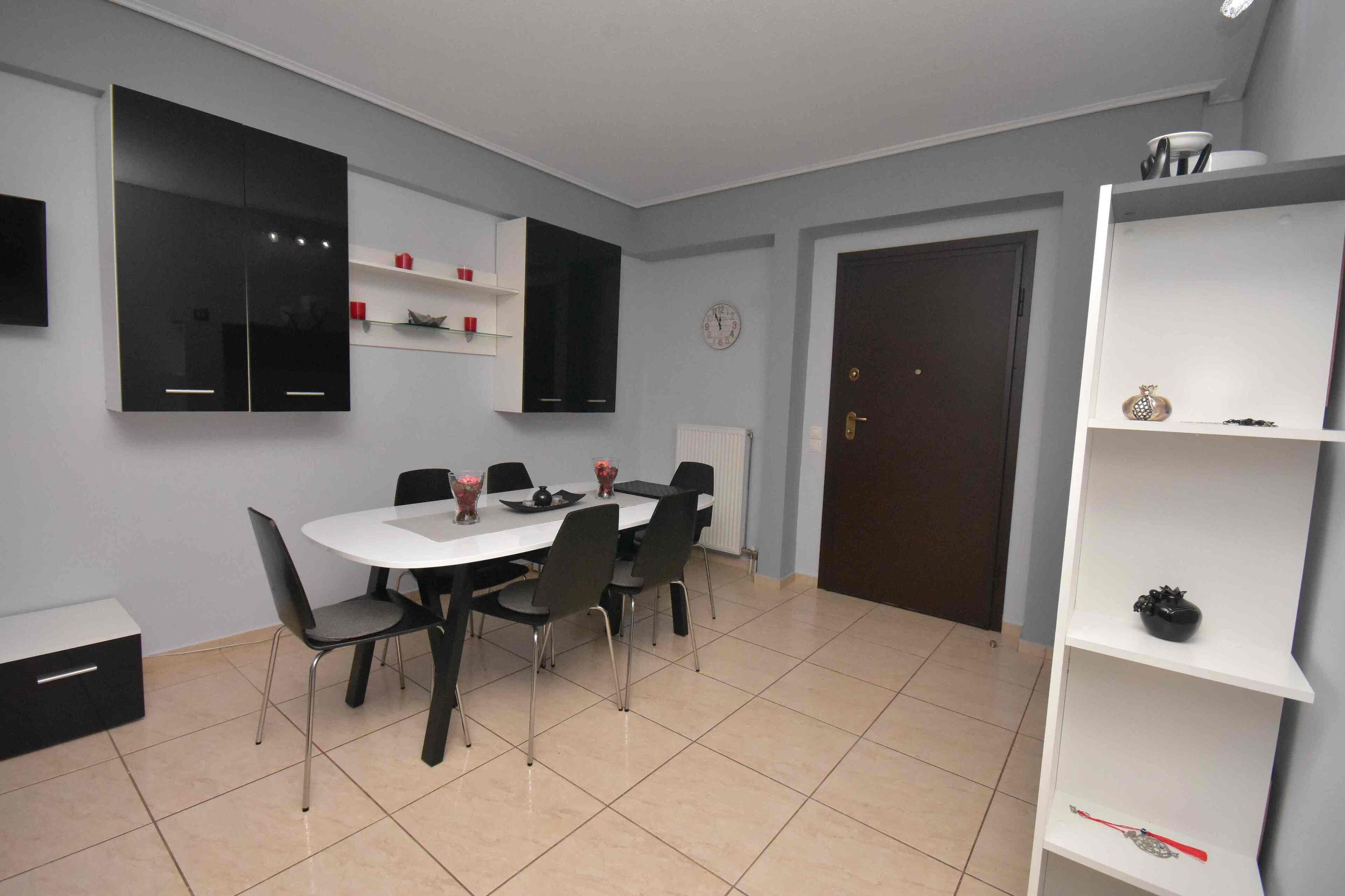 Un coin repas avec une table blanche et des chaises noires, un meuble noir placé au-dessus de la table et une étagère blanche ornée d’objets décoratifs.