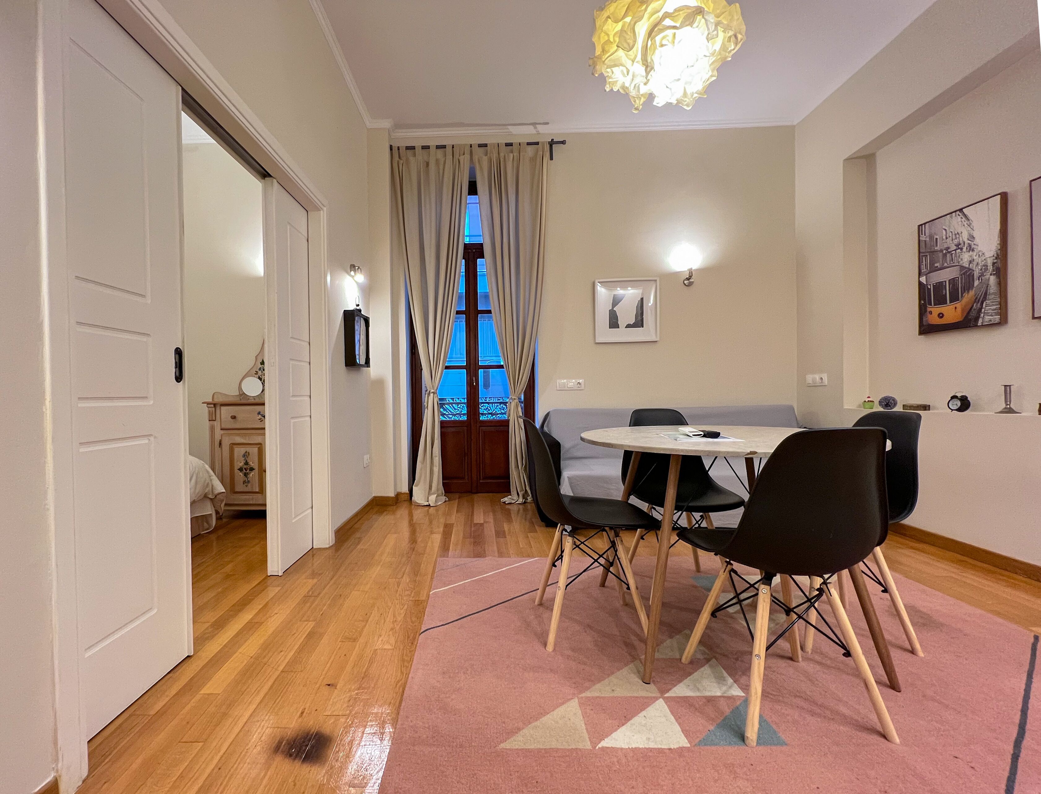 A dining area with a round table, four chairs, a pink rug, and a view of a balcony through a window with curtains.