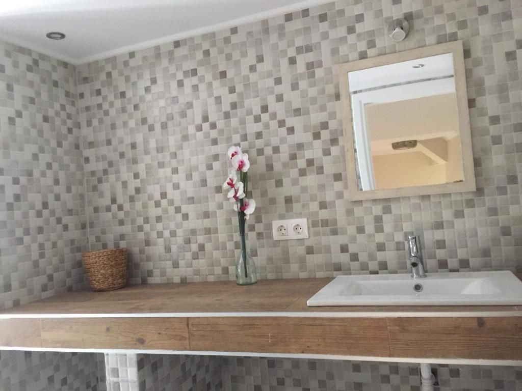 Un baño con pared de azulejos, tocador de madera, lavamanos blanco, espejo y un florero con flores.