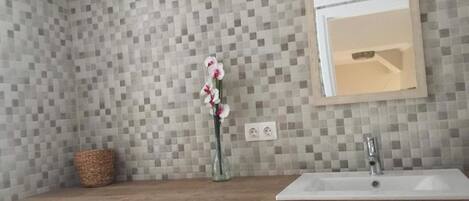 Un baño con pared de azulejos, tocador de madera, lavamanos blanco, espejo y un florero con flores.