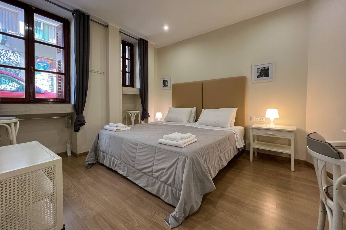 Ein Hotelzimmer mit einem großen Bett, zwei Nachttischen, einem Sessel, einem kleinen Tisch und einem Fenster mit Vorhängen.
