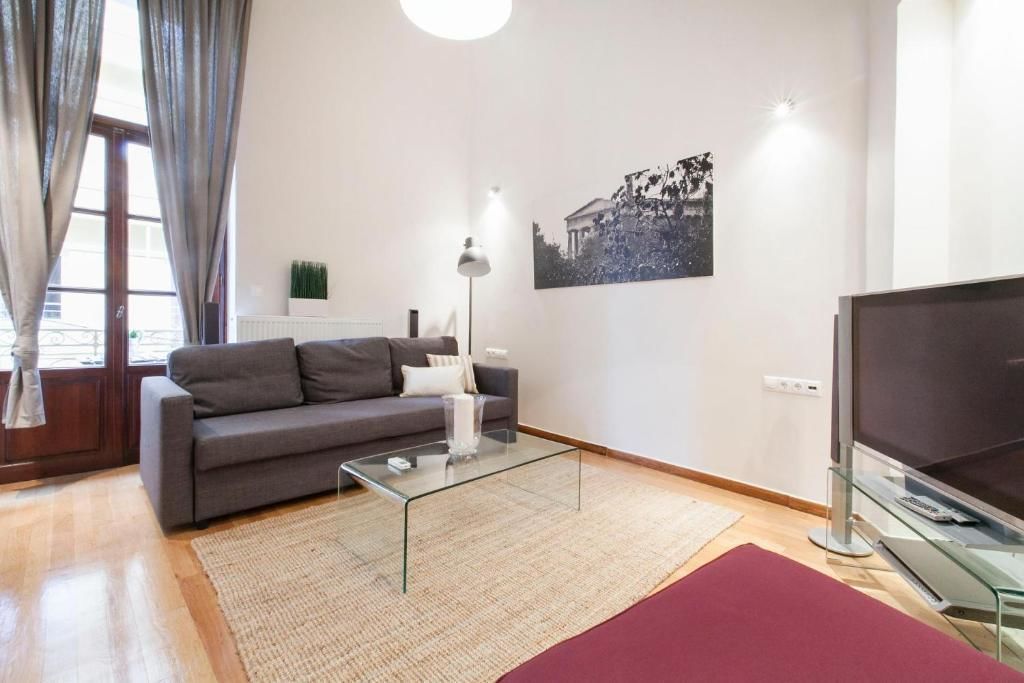 A modern living room with a grey sofa, a glass coffee table, a flat-screen TV, and a large window with curtains.